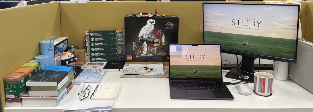 My desk in the lab: LEGO Hedwig, a stack of Nespresso pods, books, notebooks, and two screens playing the same classical-music study video.
